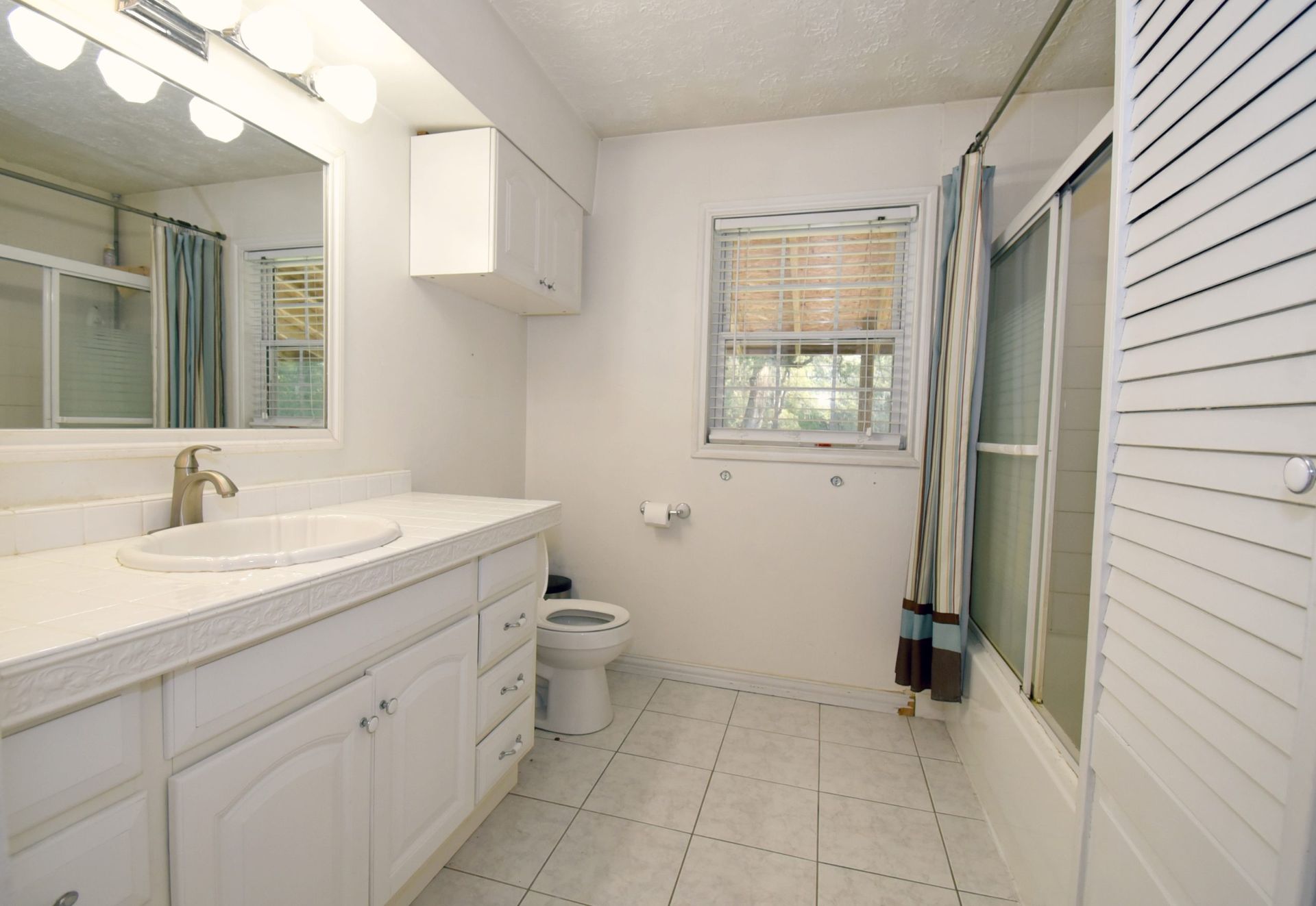 White bathroom with vanity, toilet, shower, and window with blinds.