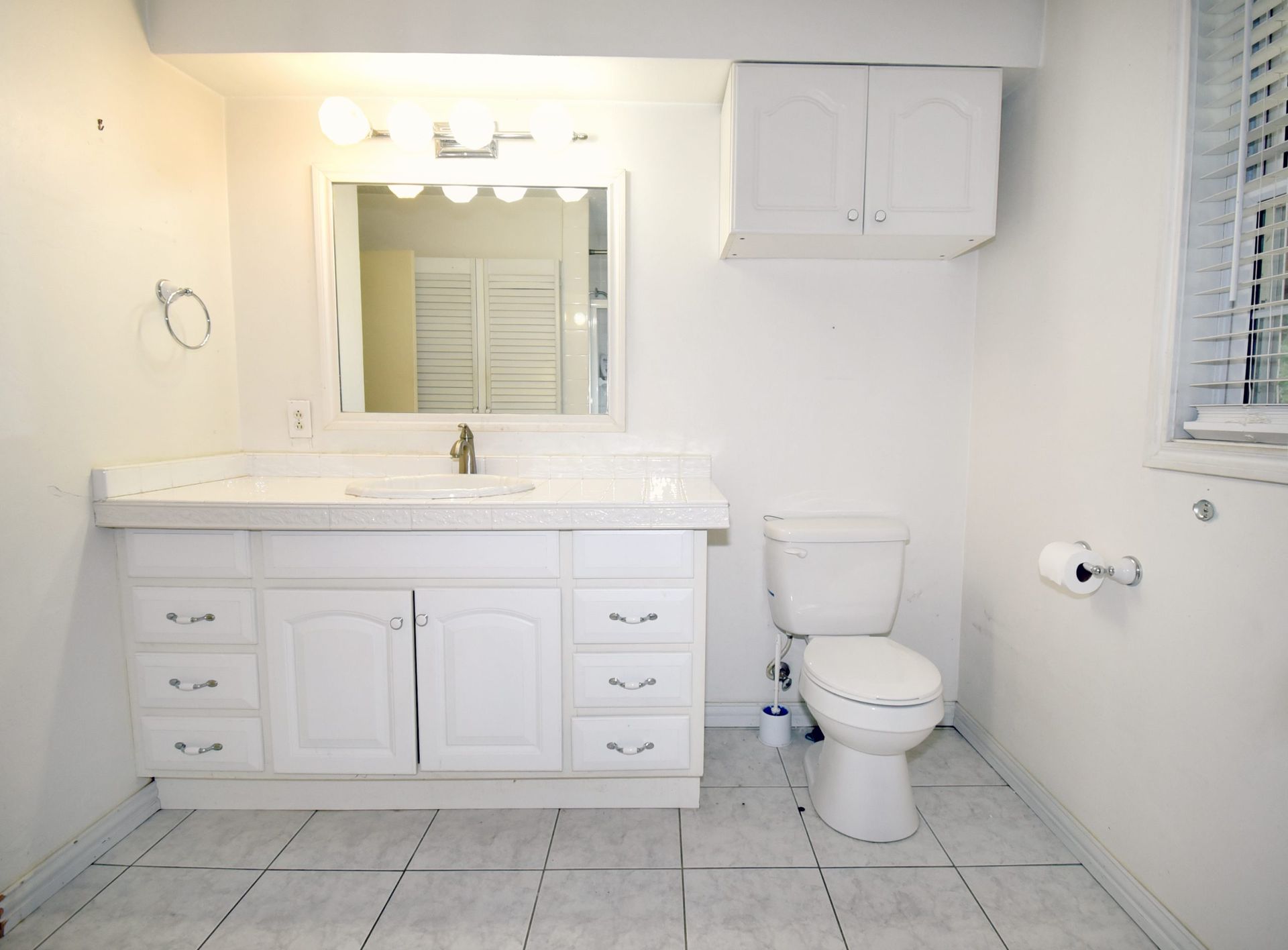 White bathroom with vanity, toilet, and overhead cabinet. Tile floor, window with blinds.