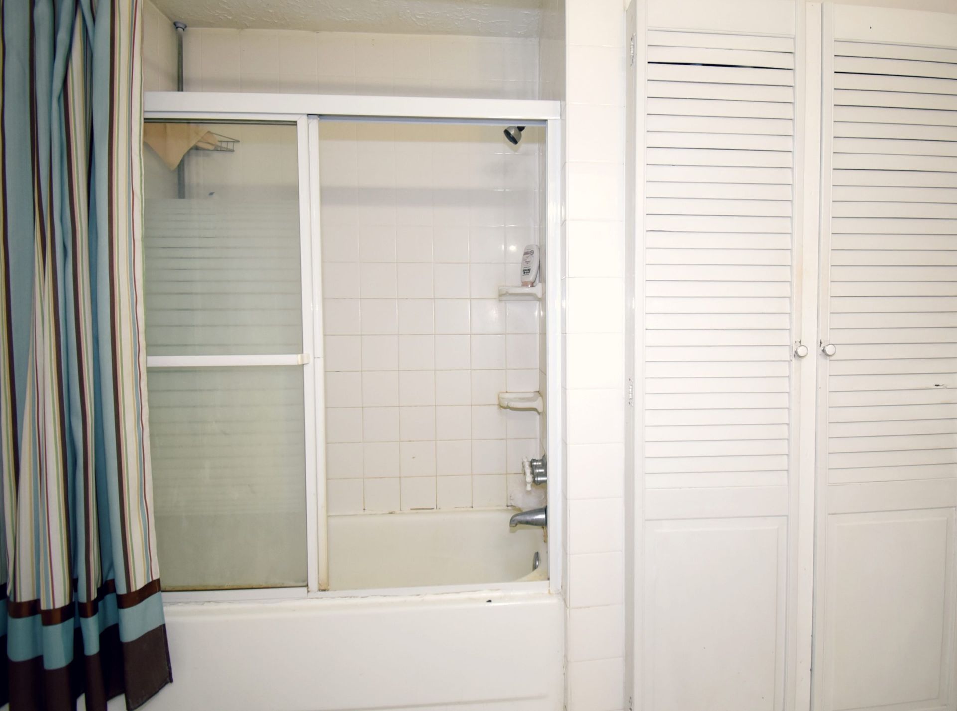 Bathroom with a shower/tub combo, sliding glass door, blue-striped shower curtain, and white shutter doors.