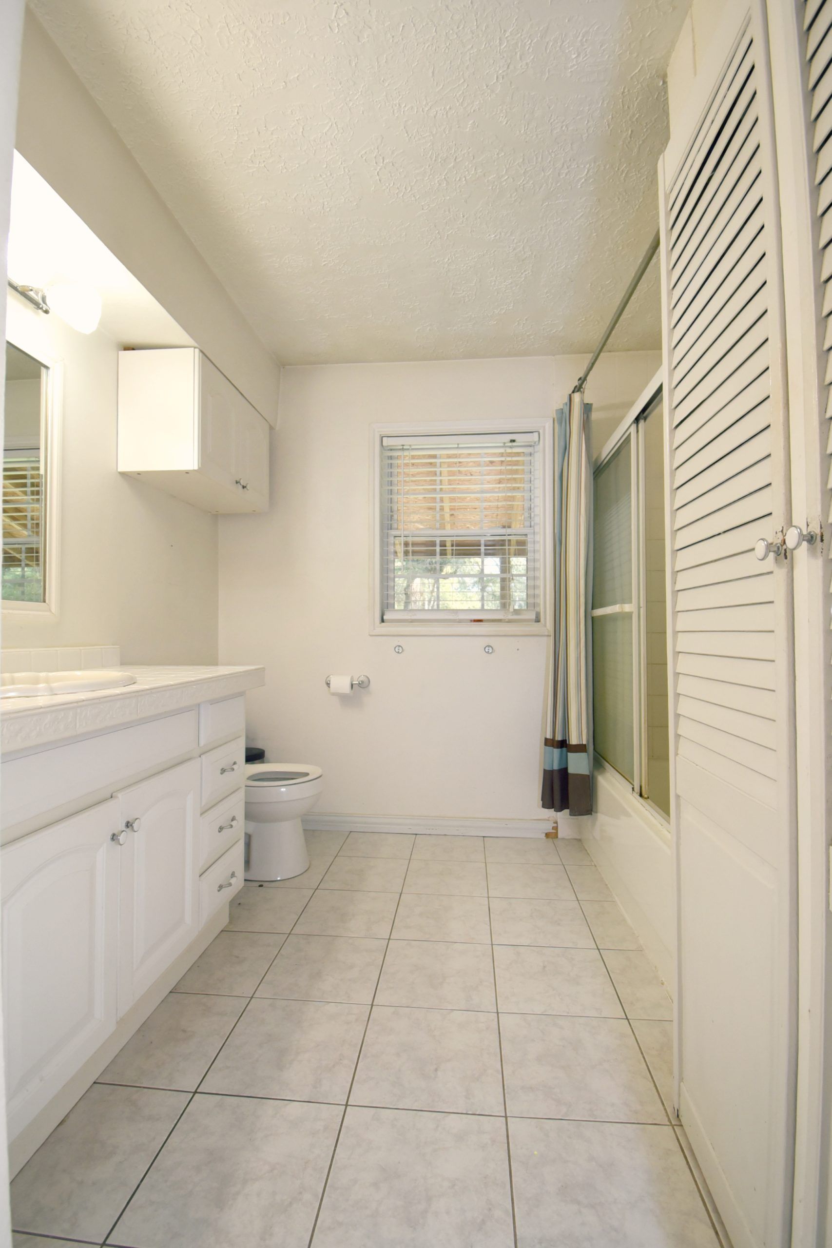 White bathroom with a vanity, toilet, and shower. A window is in the center.