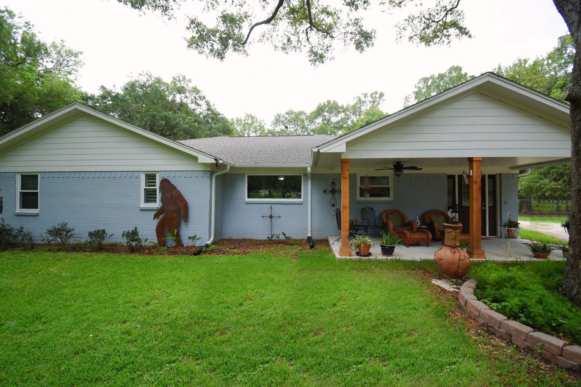 A light blue ranch-style house with Bigfoot art on the side and a covered porch, set on a grassy lawn.