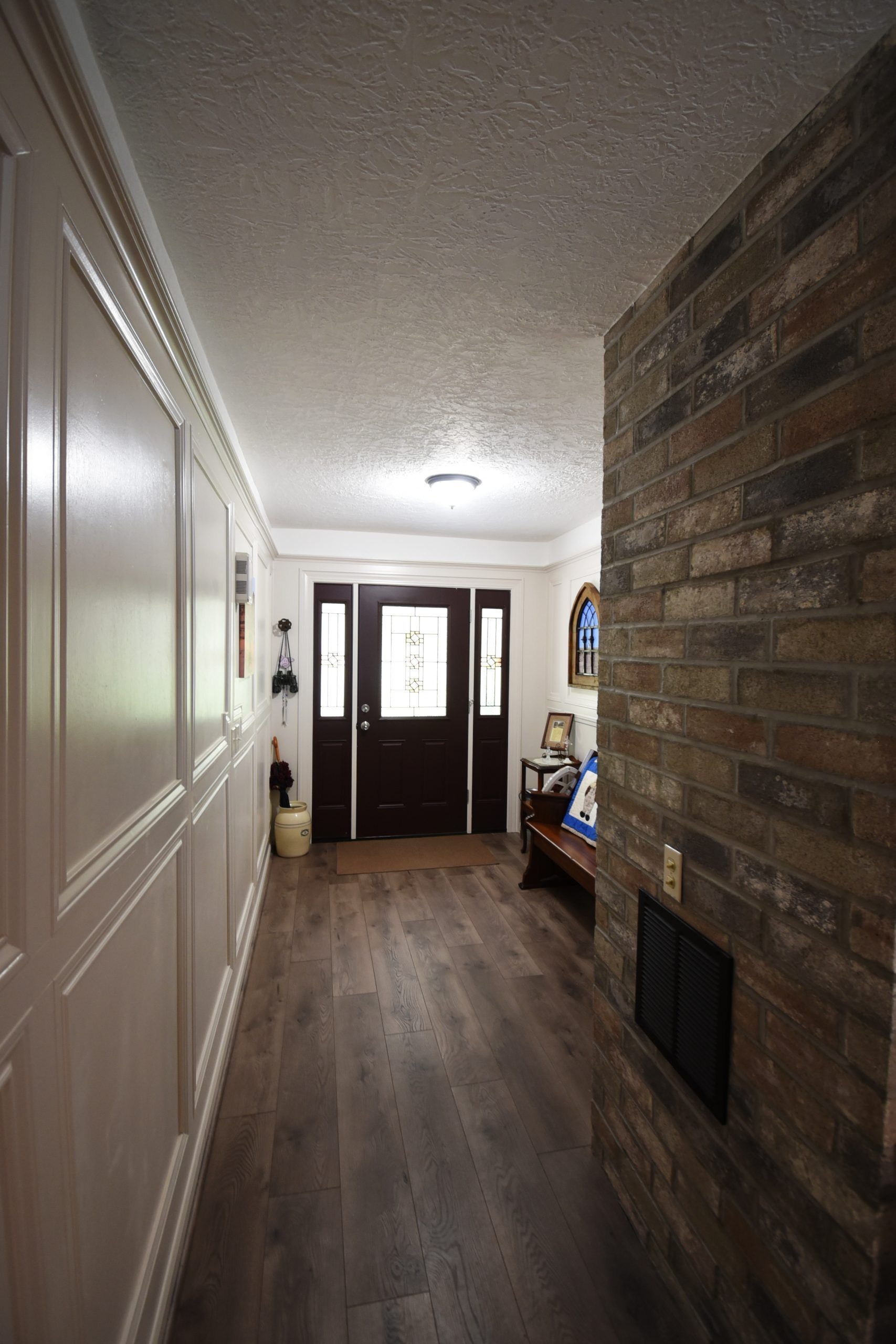 Hallway with wood floor and white paneled wall, leading to a dark wood door with sidelights; brick wall on the right.
