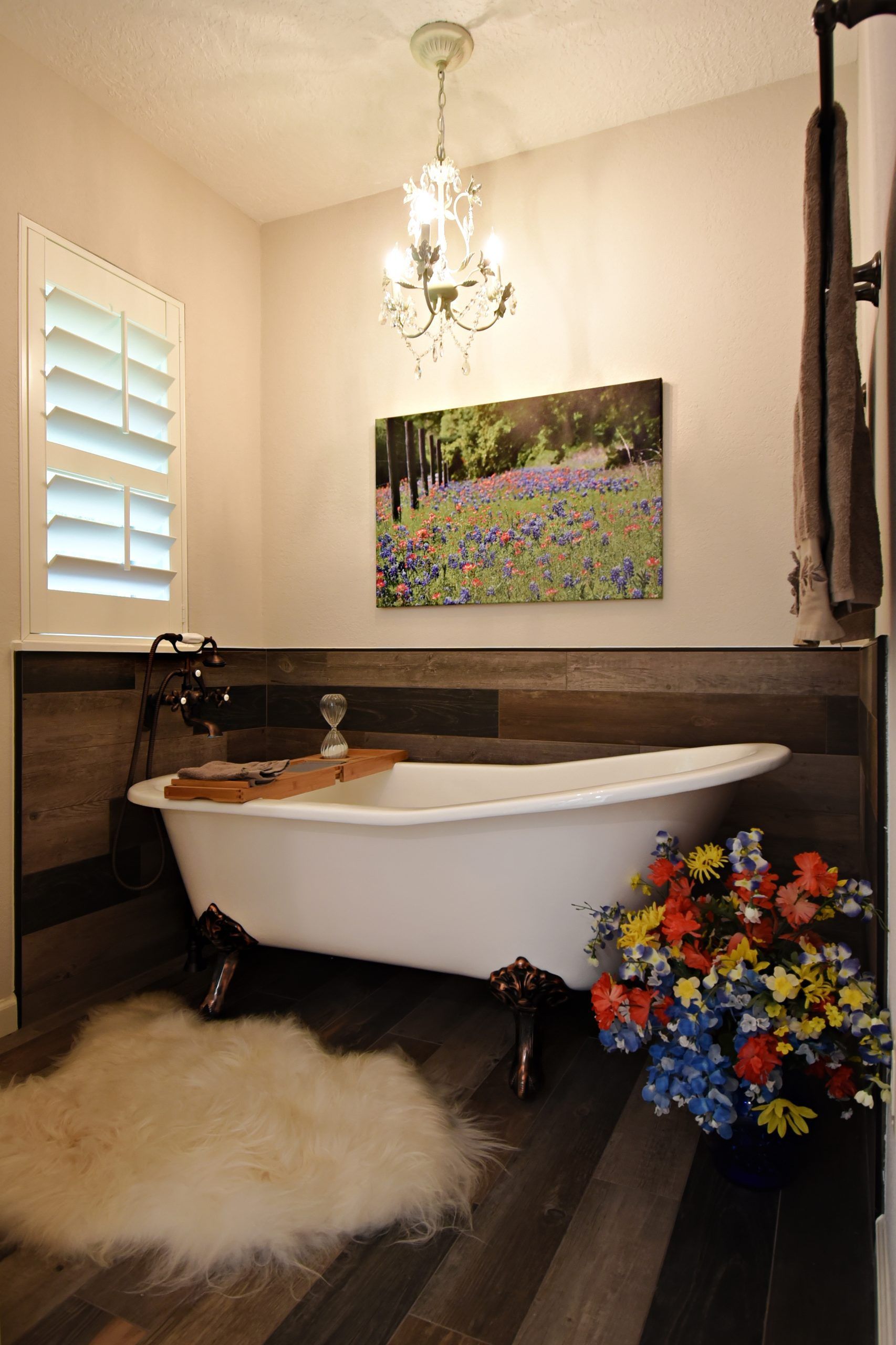 Claw-foot bathtub in rustic bathroom with wood floors, shutters, and floral artwork; chandelier and flowers.