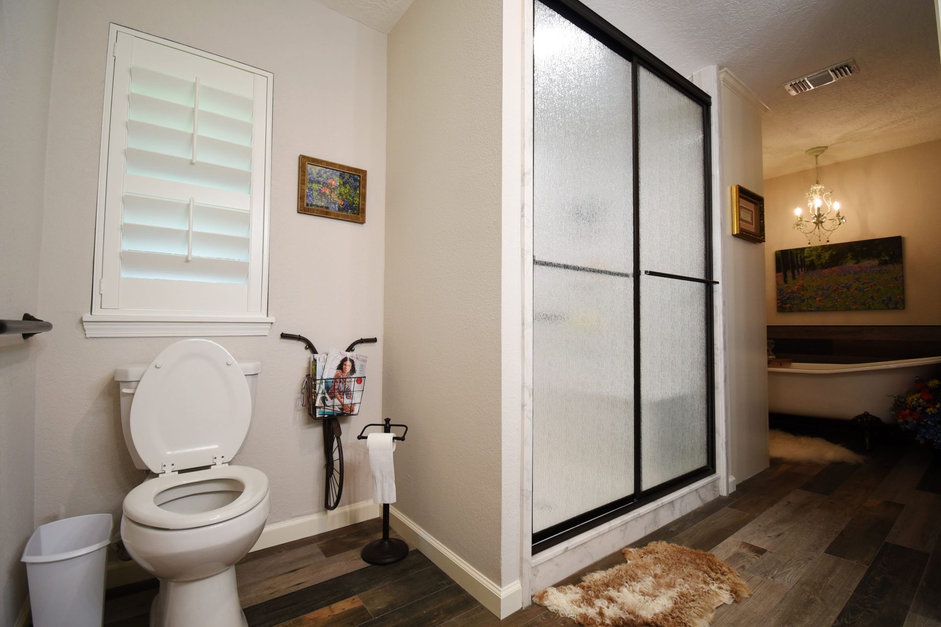 Bathroom with a toilet, shower, and window with shutters; wooden floor and a rug.