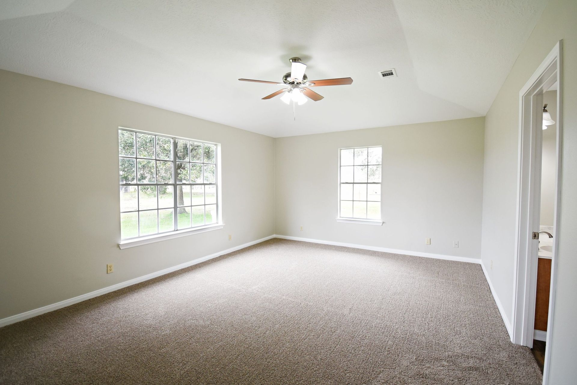 Empty bedroom with beige walls, brown carpet, two windows, and a ceiling fan.