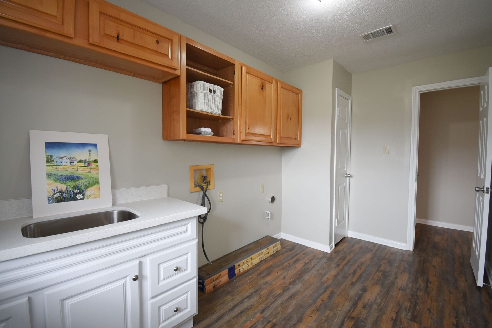Laundry room with cabinets, a sink, and a doorway to another room. Brown wood floors, light grey walls.