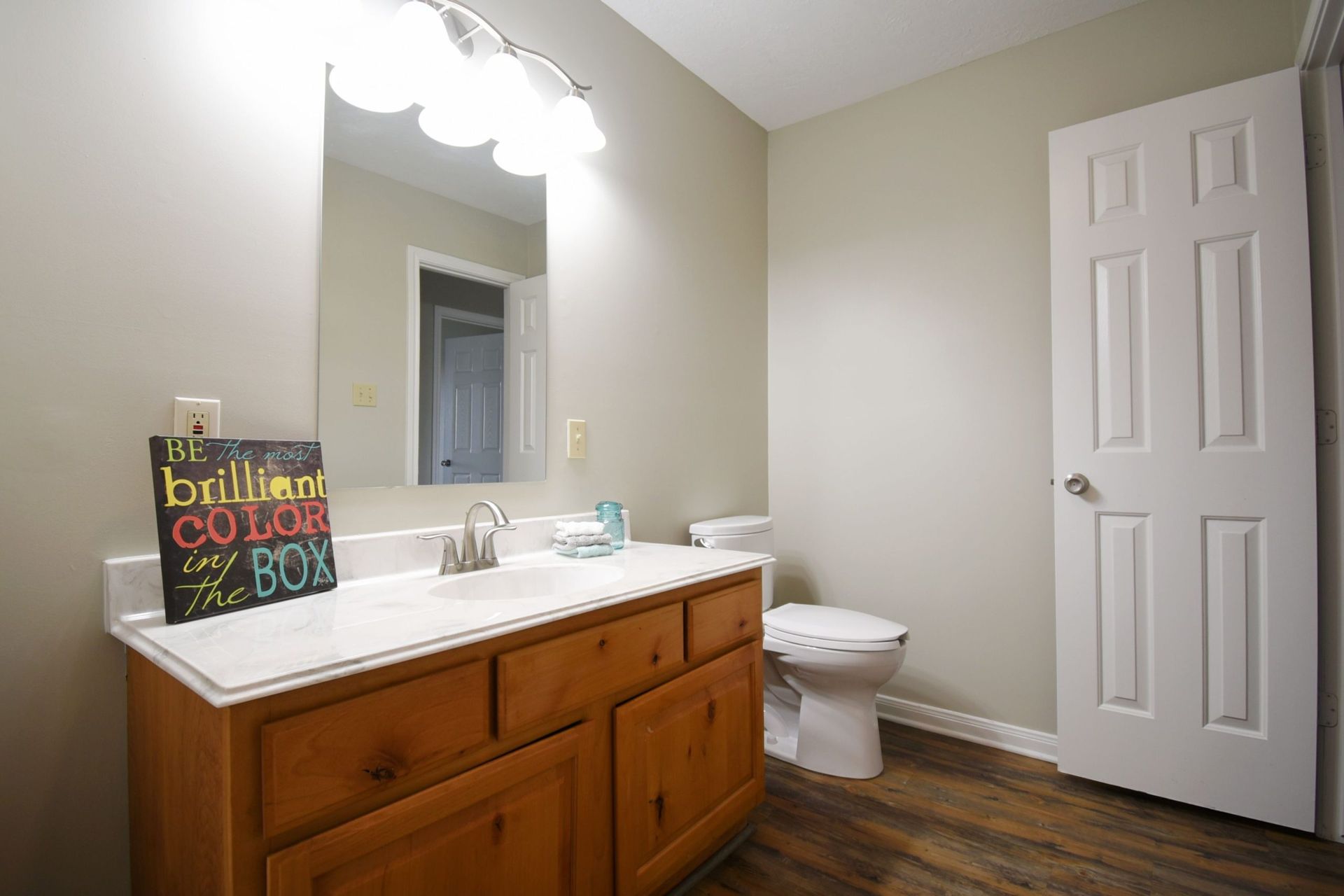 Bathroom with light-colored walls, wooden vanity, toilet, and white door. A mirror and a sign are above the sink.