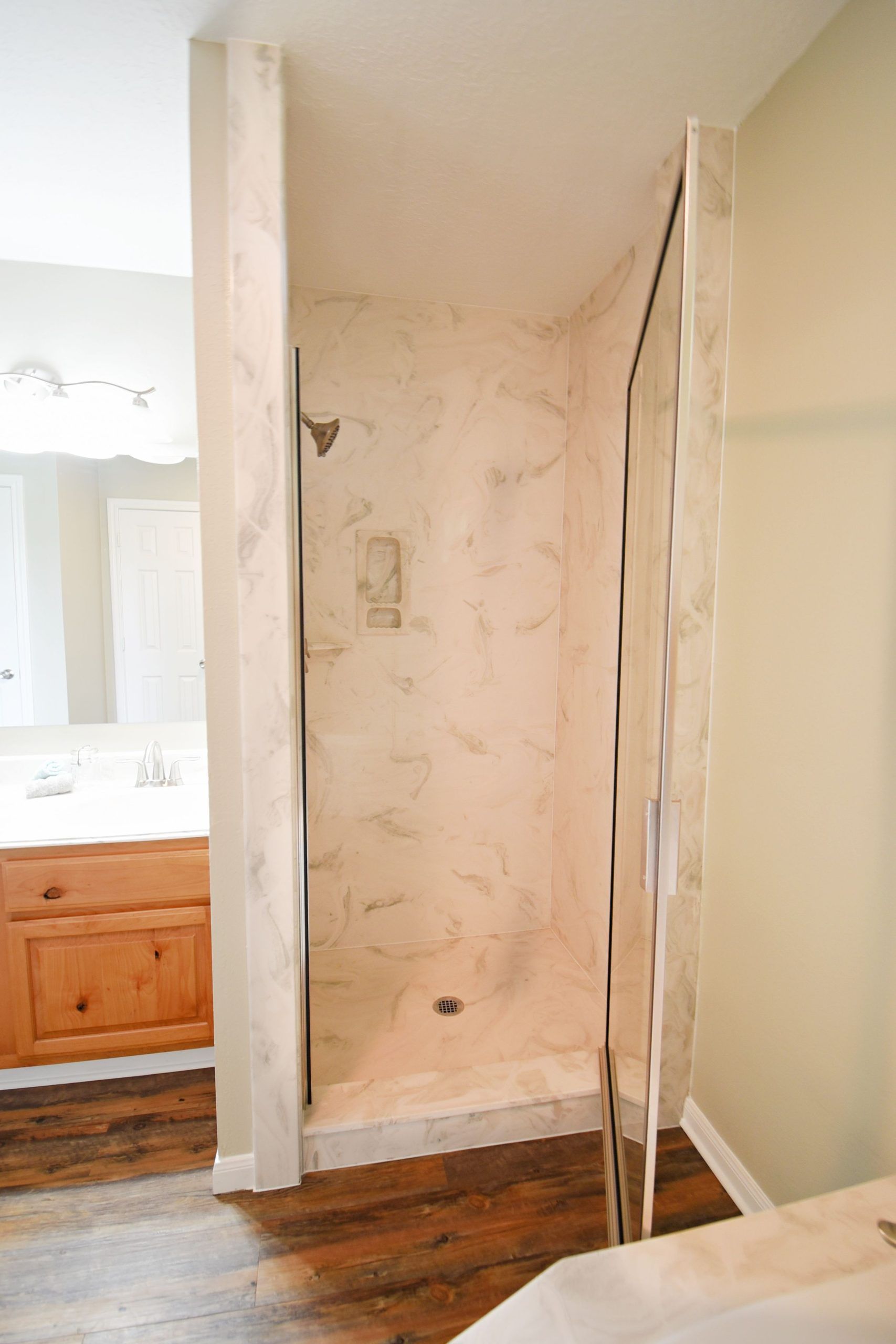 Shower stall with marble-like walls. Wooden floor in front. Beige walls in the bathroom, wood vanity visible.