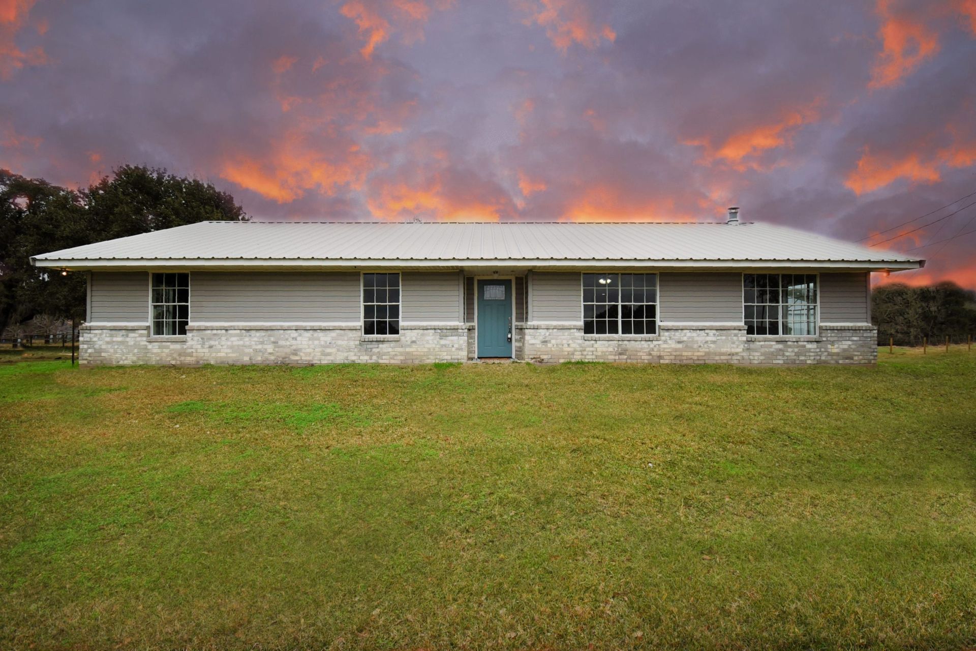 Ranch house with gray siding, stone base, and teal door on a grassy field under a dramatic sunset.