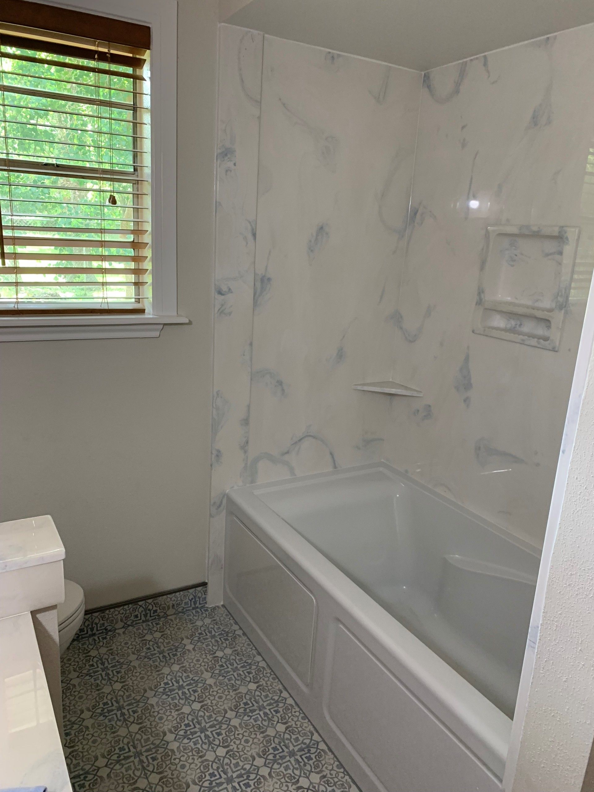 Bathroom with white tub, marbled blue and white walls, and a window with blinds.