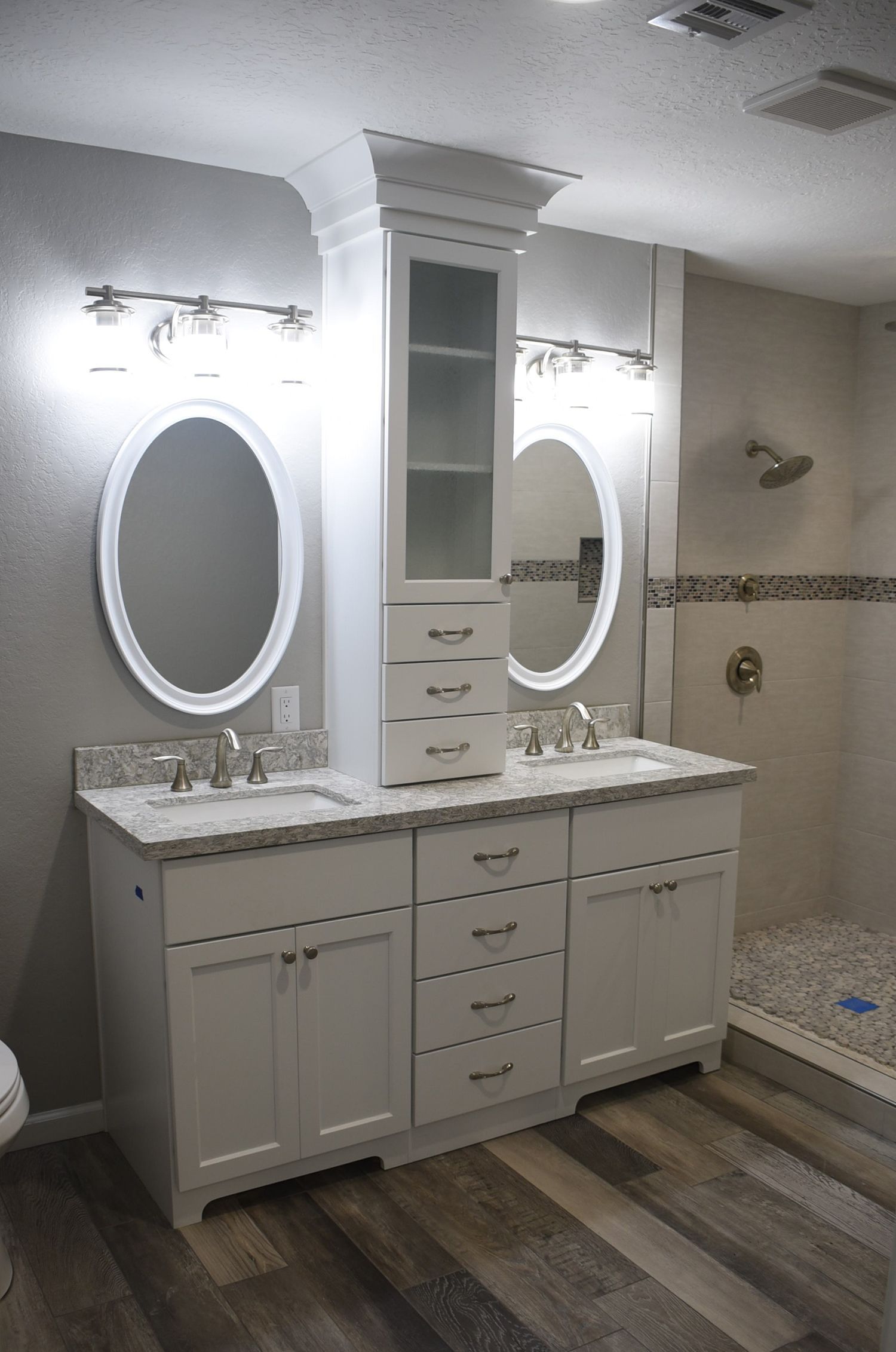 Bathroom with white vanity, oval mirrors, and a glass-doored cabinet between two sinks.