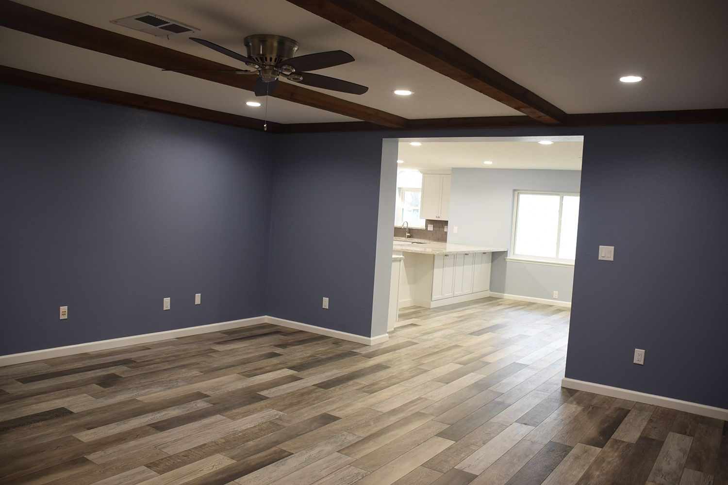 Open living room with blue walls, wood beams, and wood-look flooring, opening to white kitchen.