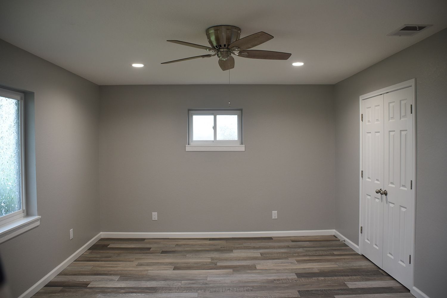 Empty room with gray walls, wood-look floor, ceiling fan, small window, and white door.