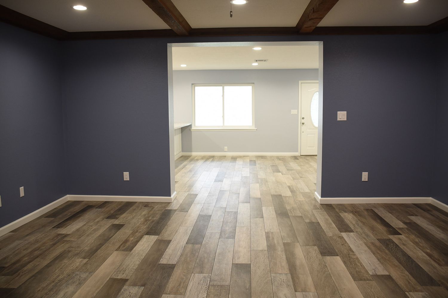 Room with blue walls, wooden beams, and hardwood floors, leading to another room with a window.