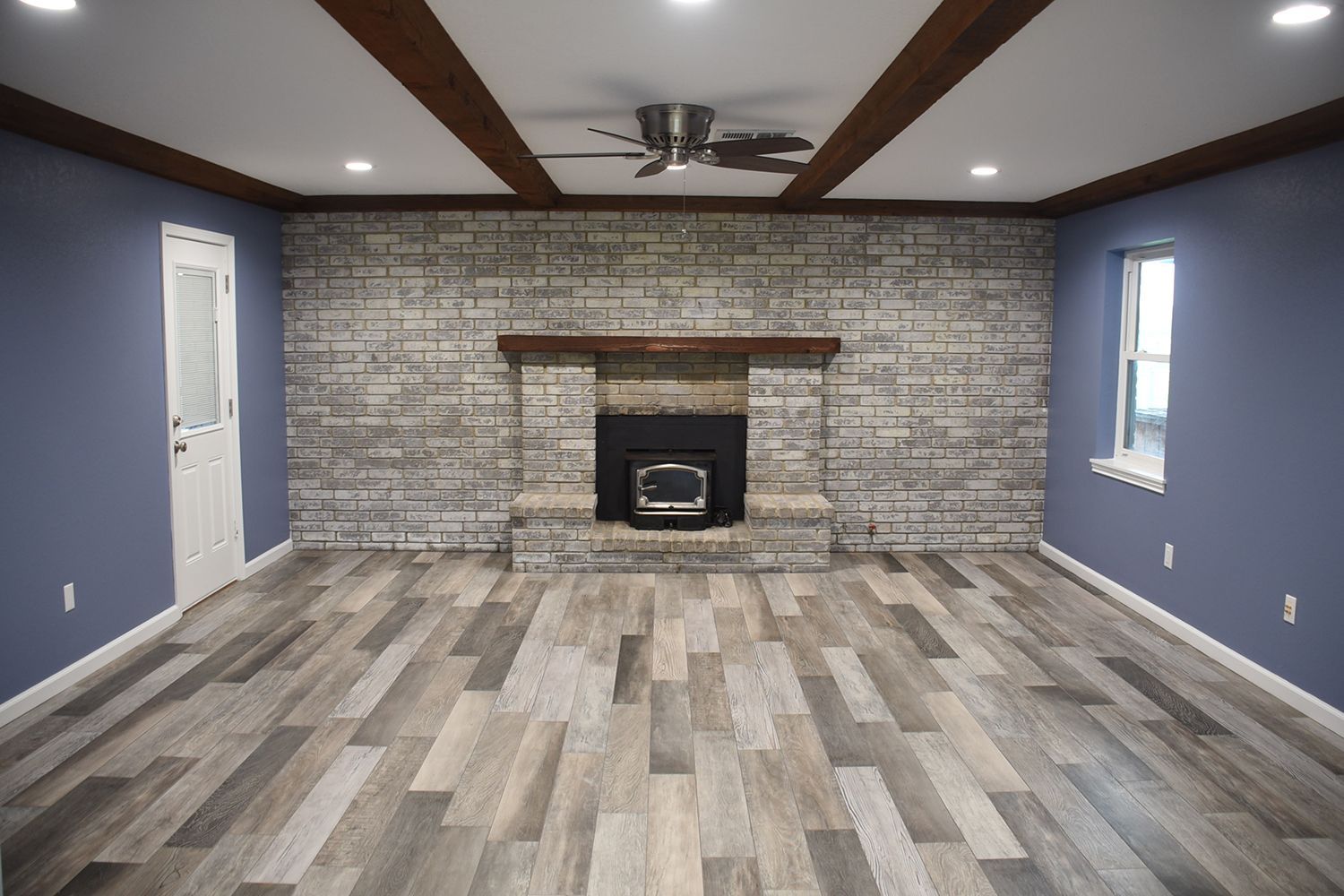 Living room with brick fireplace, wood beams, blue walls, and wood-look flooring.
