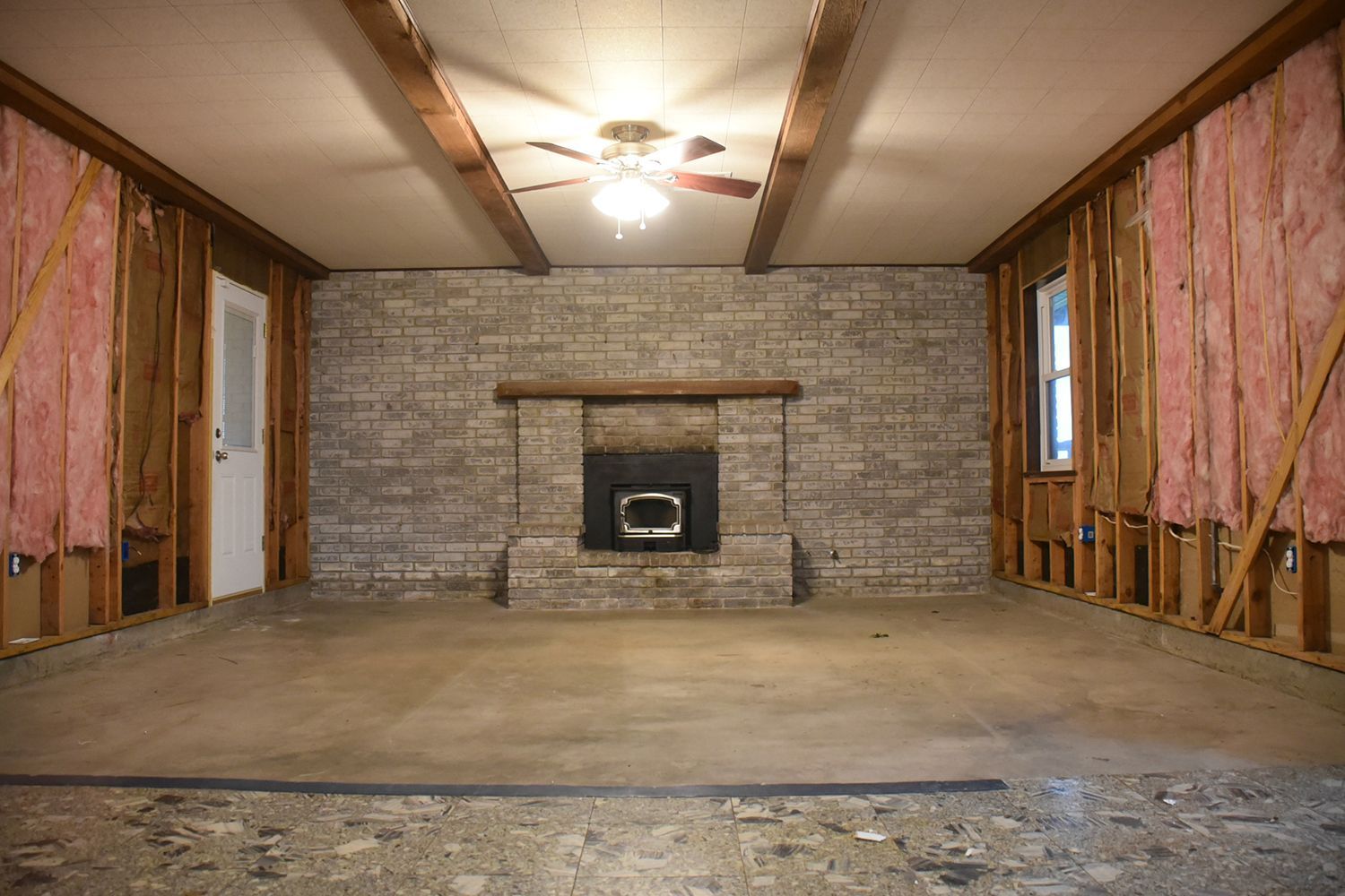Room gutted during renovation, brick fireplace, exposed framing, insulation, ceiling beams.