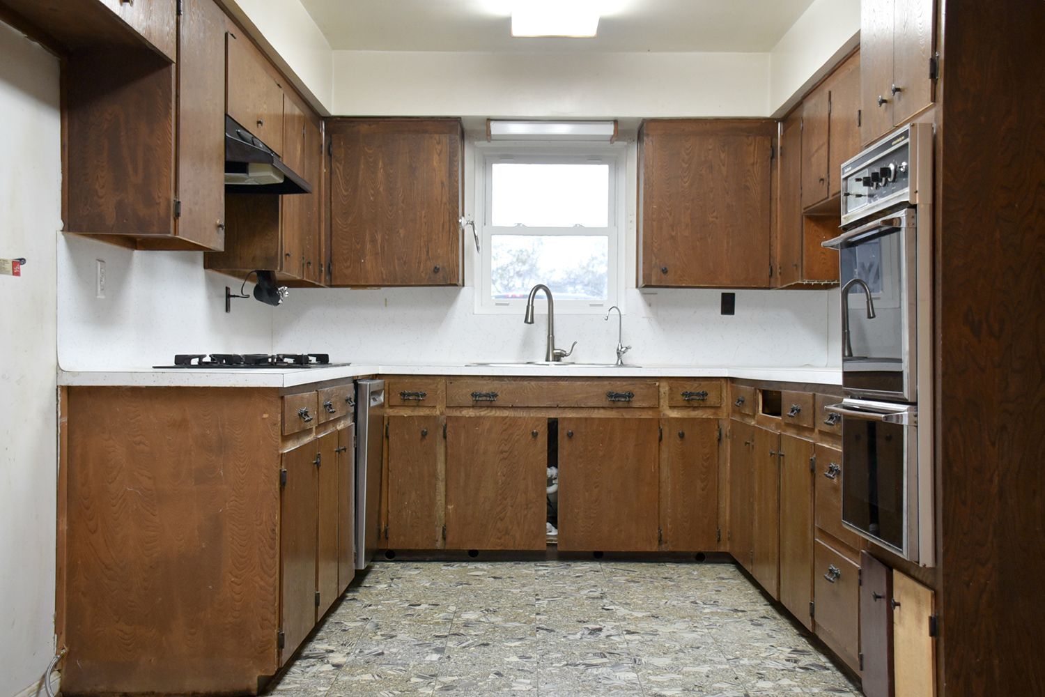 A drab, outdated kitchen with dark wood cabinets, a sink by a window, and a double oven.