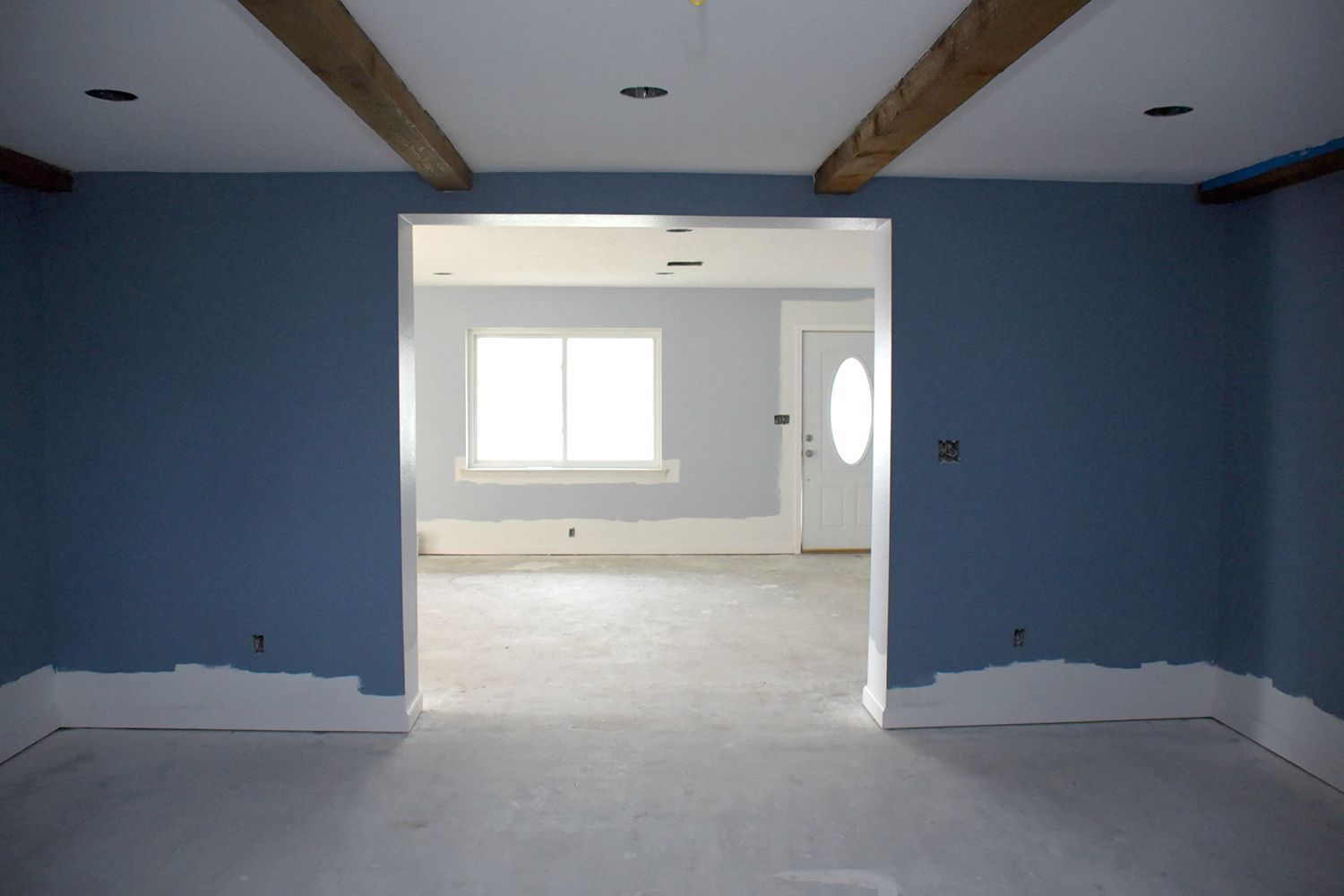 Blue-painted room with white trim and beams, leading to a brighter room with a door and window.