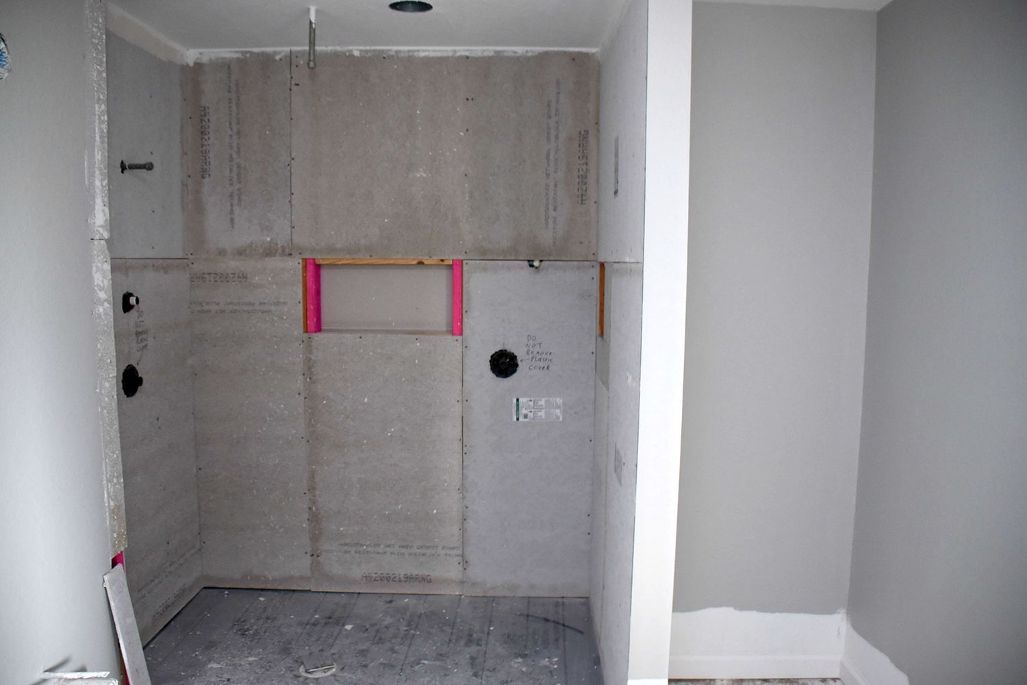 Bathroom shower under construction; gray walls, unfinished; pink window frame, black fixtures.