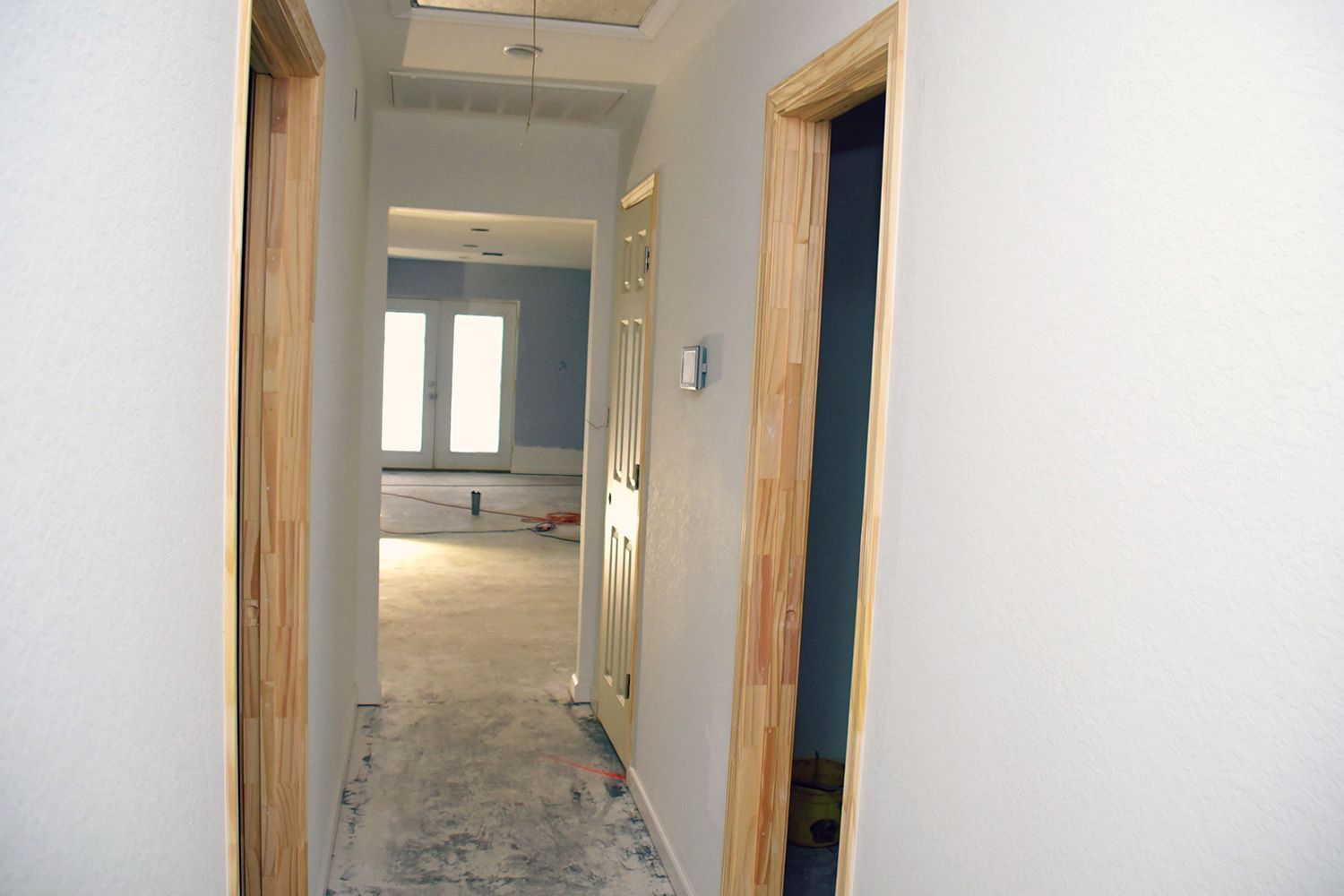Narrow hallway with doors, leading to a room with windows. White walls, wooden door frames.