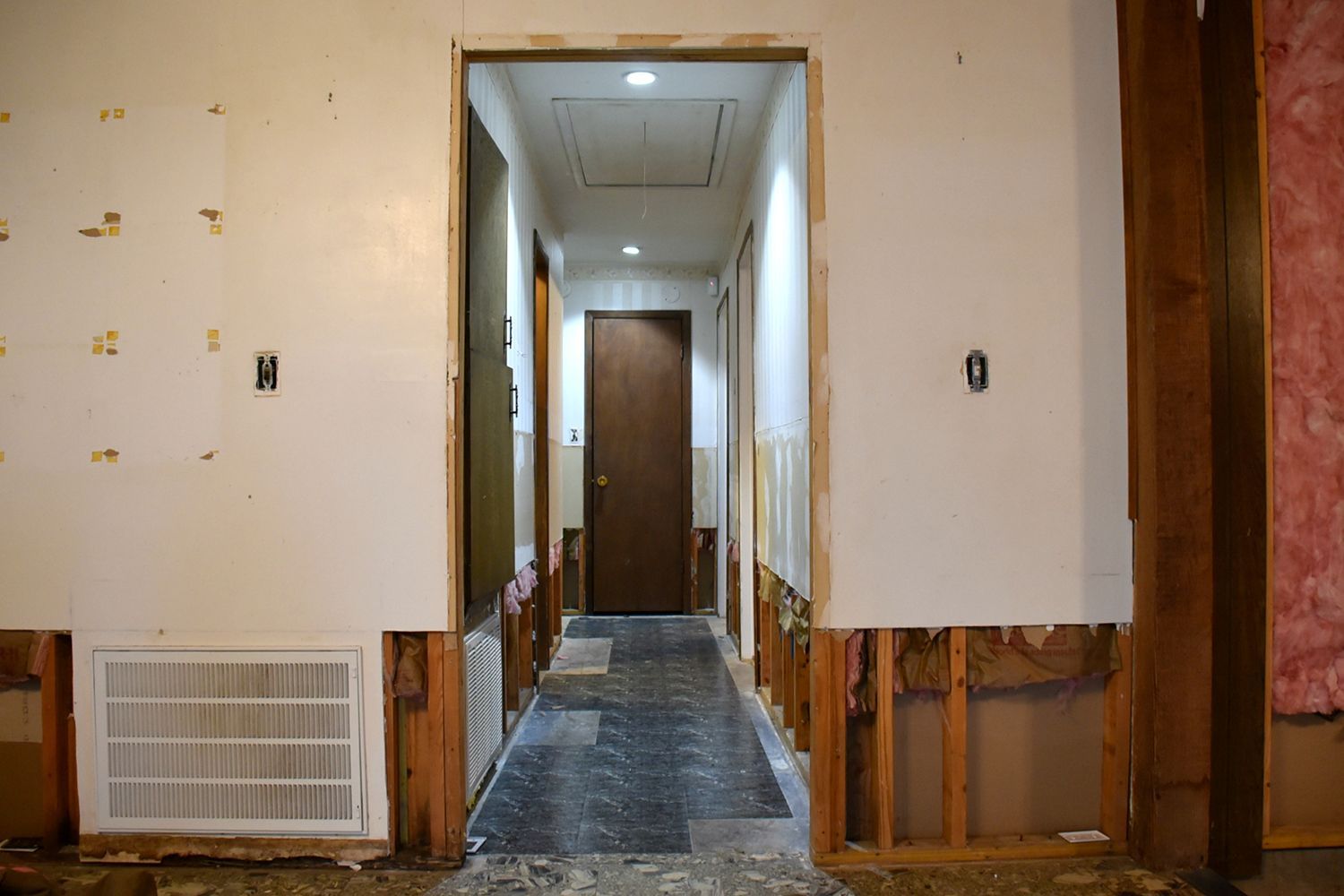Hallway under renovation; drywall removed, revealing framing and insulation; a brown door at the end.