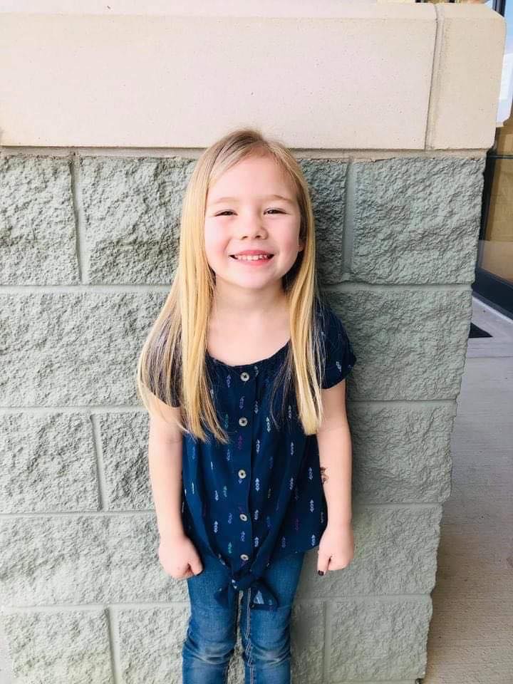 A little girl is standing in front of a brick wall and smiling.