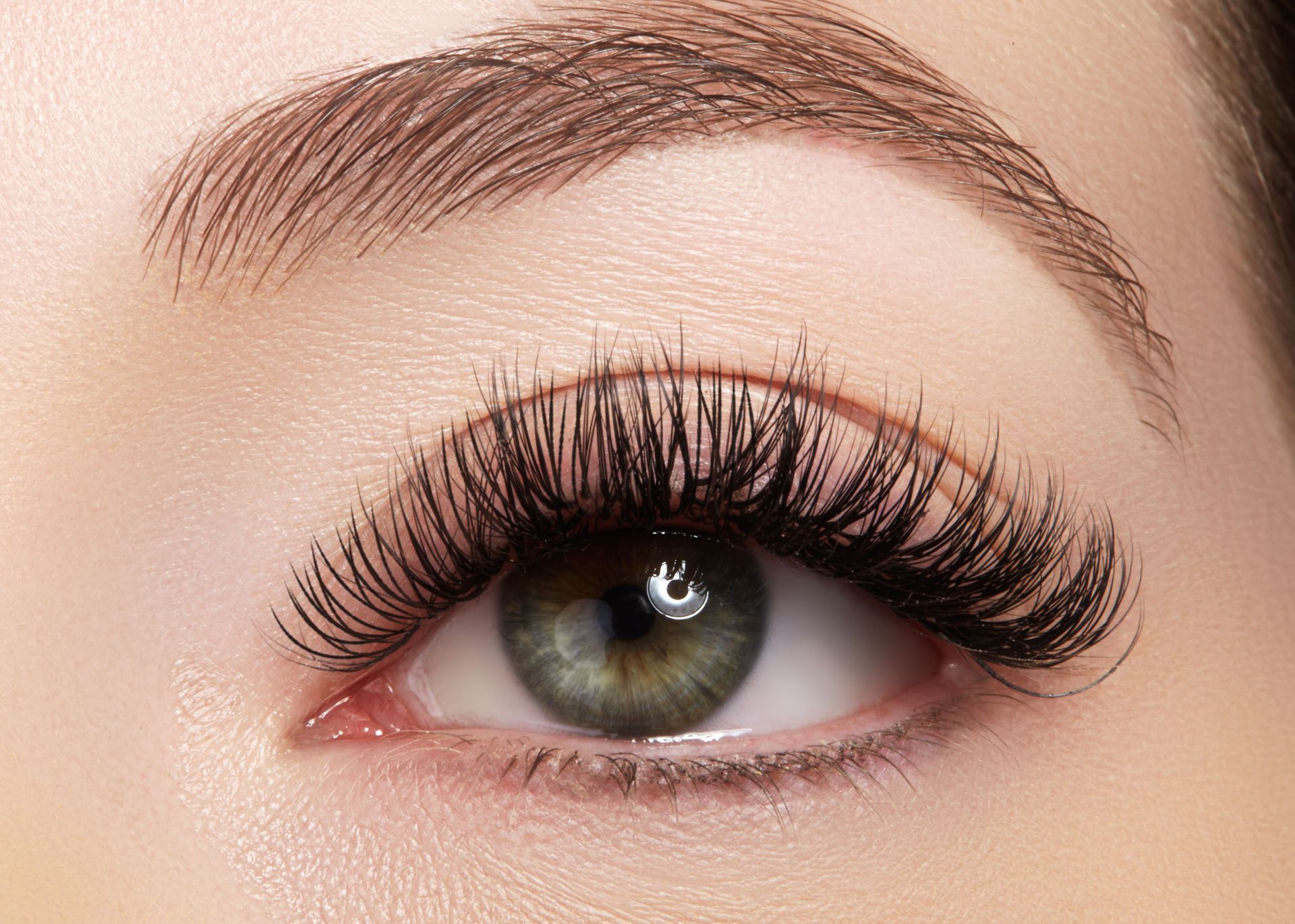 A close-up of a human eye with long, dark, voluminous eyelashes, featuring a hazel-green iris and groomed eyebrows.