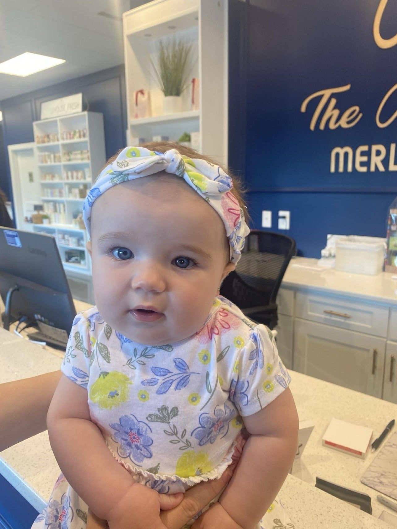 A baby girl wearing a headband is sitting at a counter in a pharmacy.