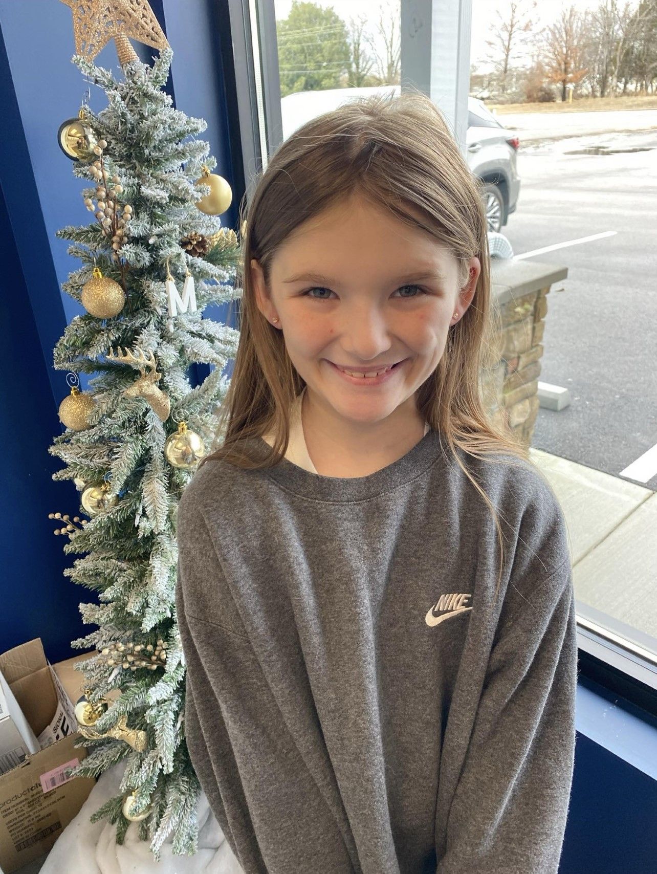 A little girl in a gray sweater is standing in front of a Christmas tree.