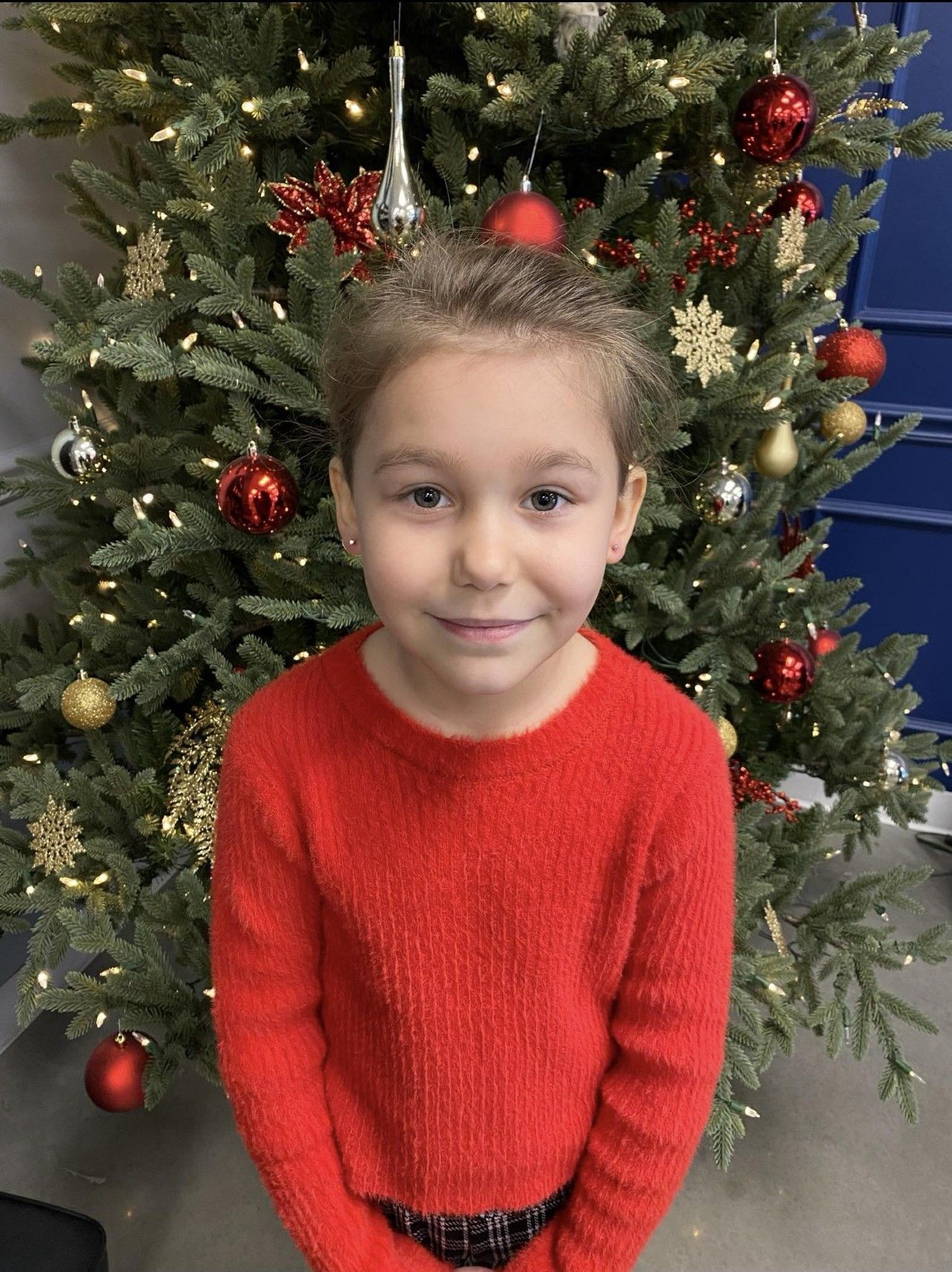 A little girl in a red sweater is standing in front of a Christmas tree.