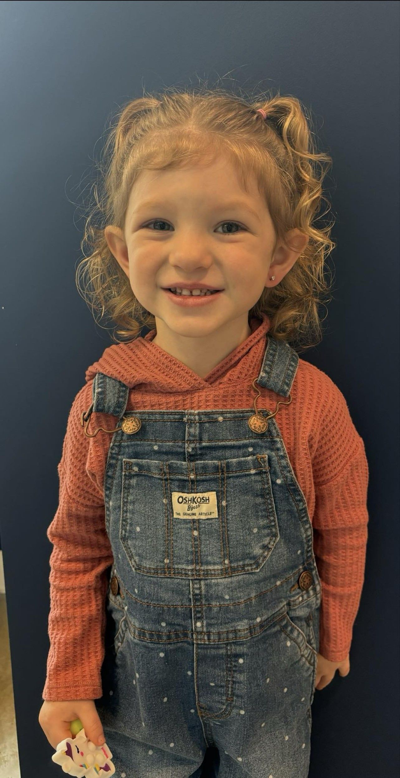 A little girl wearing overalls and a red shirt is smiling.