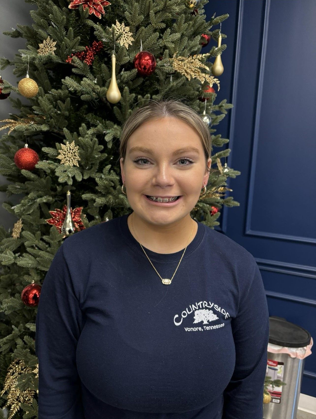 A woman in a blue shirt is standing in front of a Christmas tree.