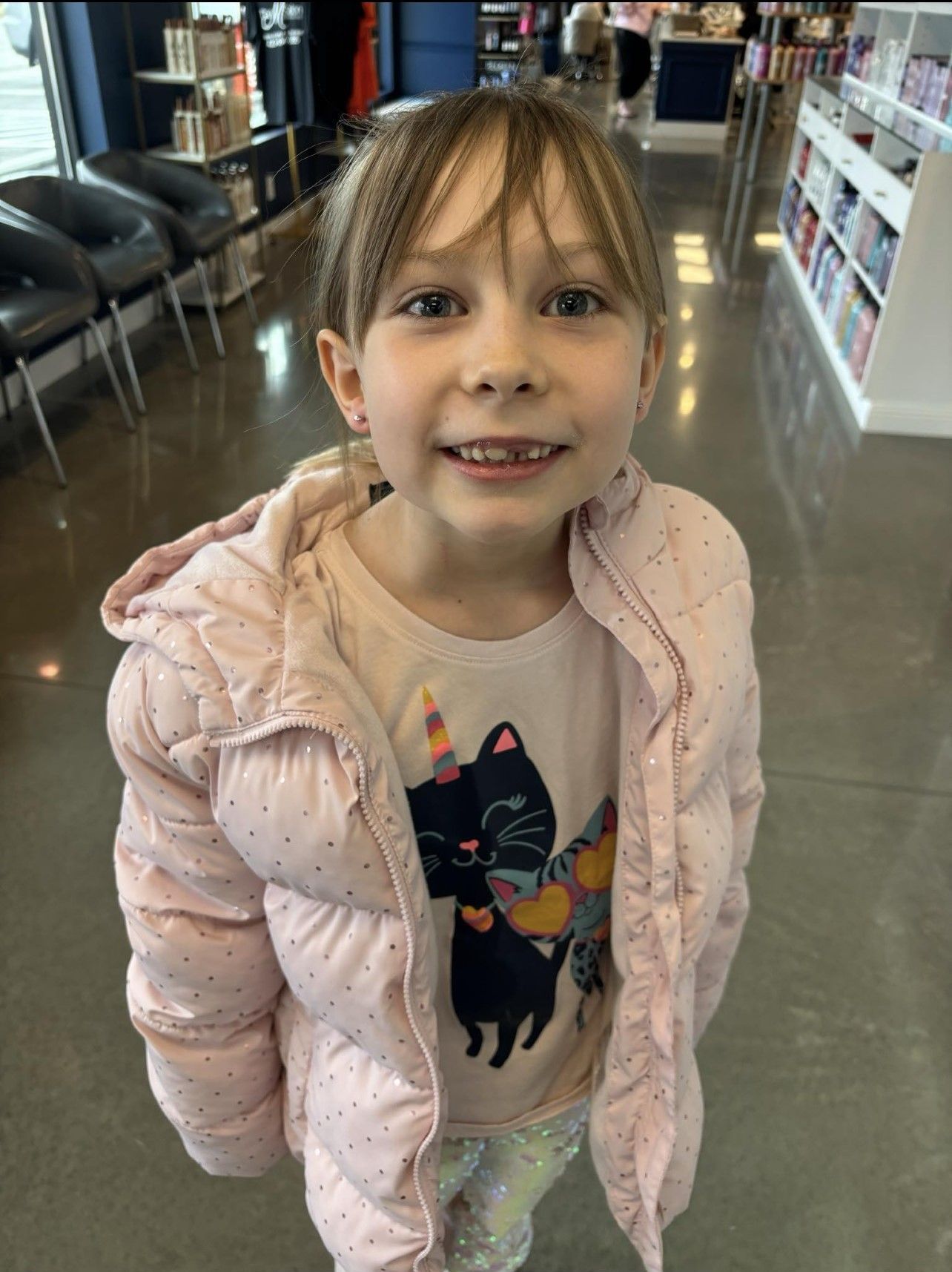 A little girl wearing a pink jacket and a cat shirt is standing in a store.
