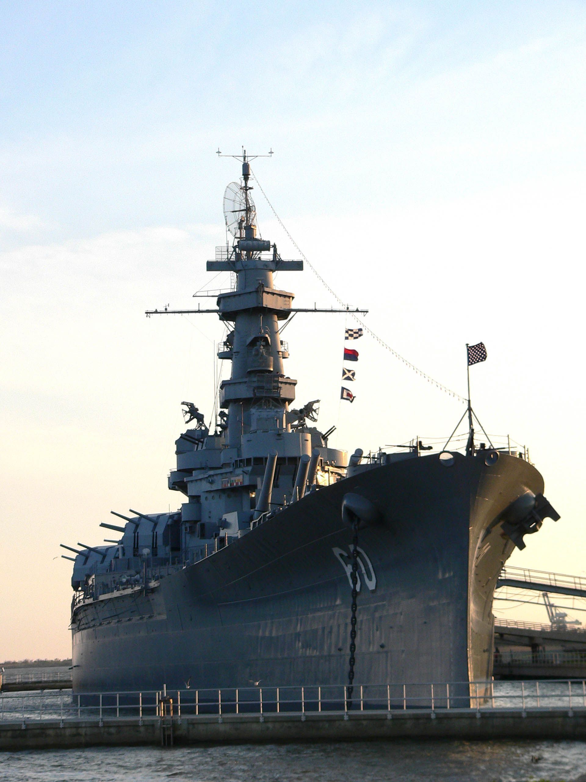 Battleship in harbor, gray hull, prominent gun turrets, flags flying, against pale sky.