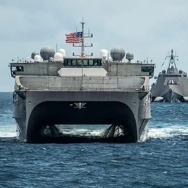 A large US Navy ship with the American flag, sailing at sea, with another ship in the background.