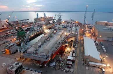 Aerial view of a naval shipyard at dusk, with multiple ships under construction and large cranes.