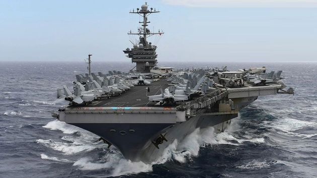 A large U.S. Navy aircraft carrier steaming through the ocean with many aircraft on deck.