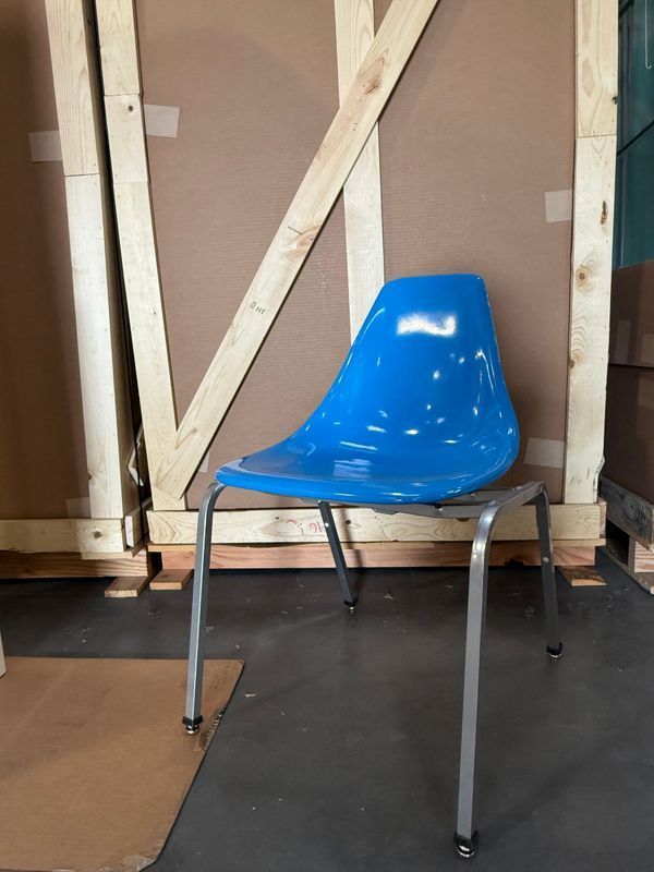 Blue plastic chair with metal legs in front of a wooden structure on a concrete floor.