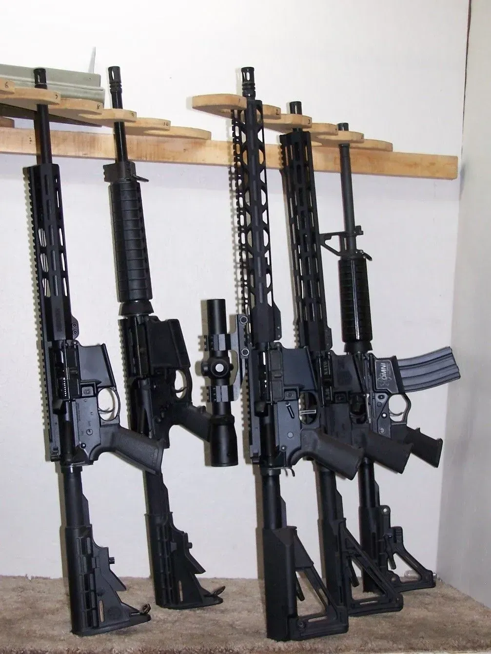 Five black rifles hanging on a wooden rack against a white wall.
