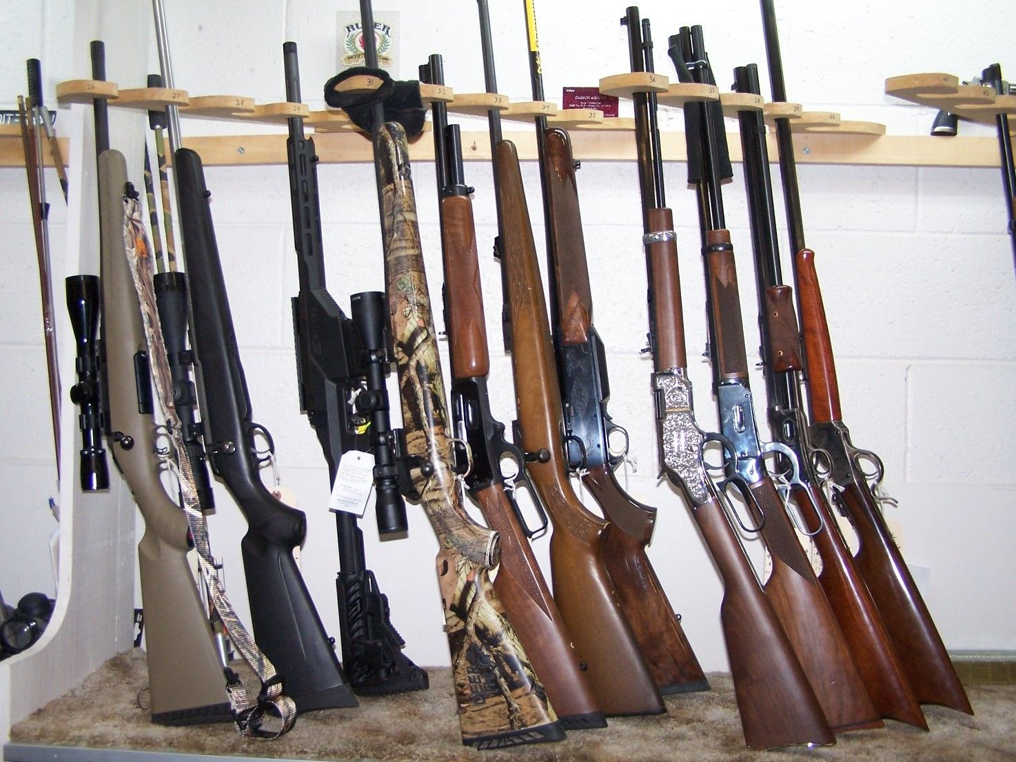 Rifles displayed on a wooden rack, varying colors and styles. Indoor setting, likely a gun shop.