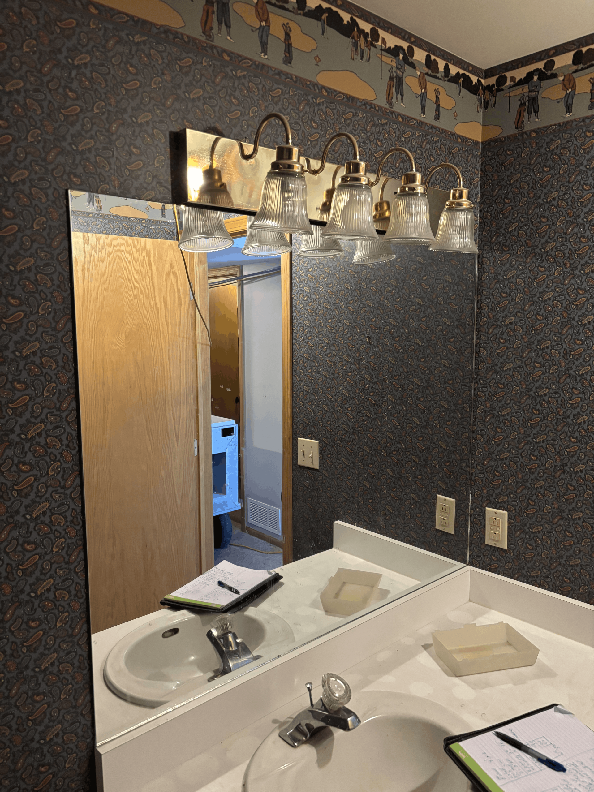 Bathroom vanity with mirror, lights, double sink, and an open door showing a laundry room.