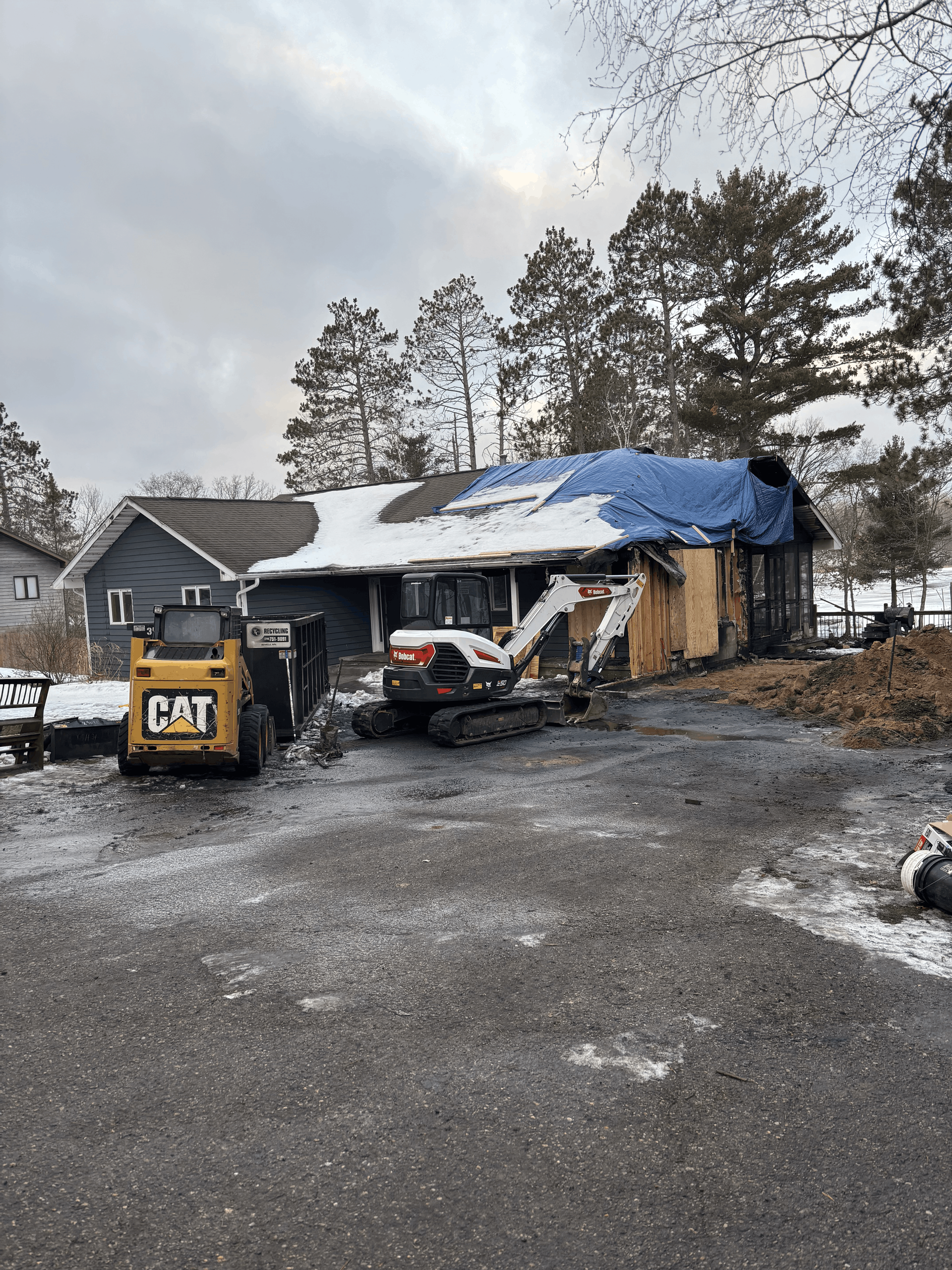Fire-damaged house with blue tarp roof, CAT loader, and debris in a snowy driveway