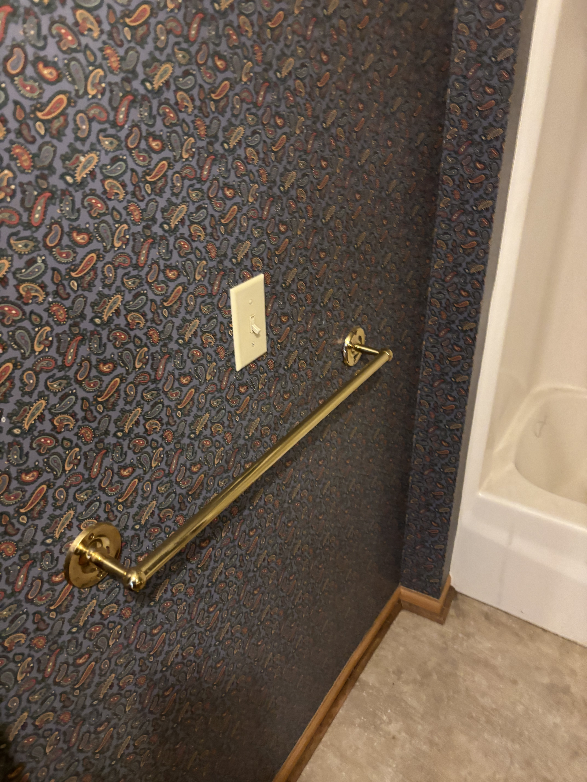Wallpapered stair landing with brass handrail, outlet, and edge of a bathtub or shower curtain.