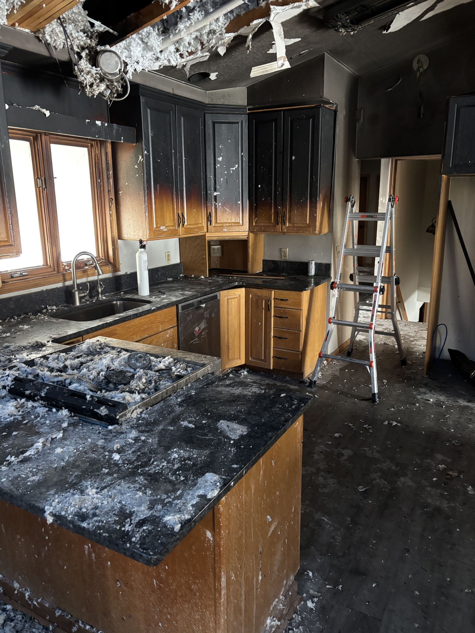 Fire-damaged kitchen with charred cabinets, blackened countertops, and a ladder amid heavy soot damage.