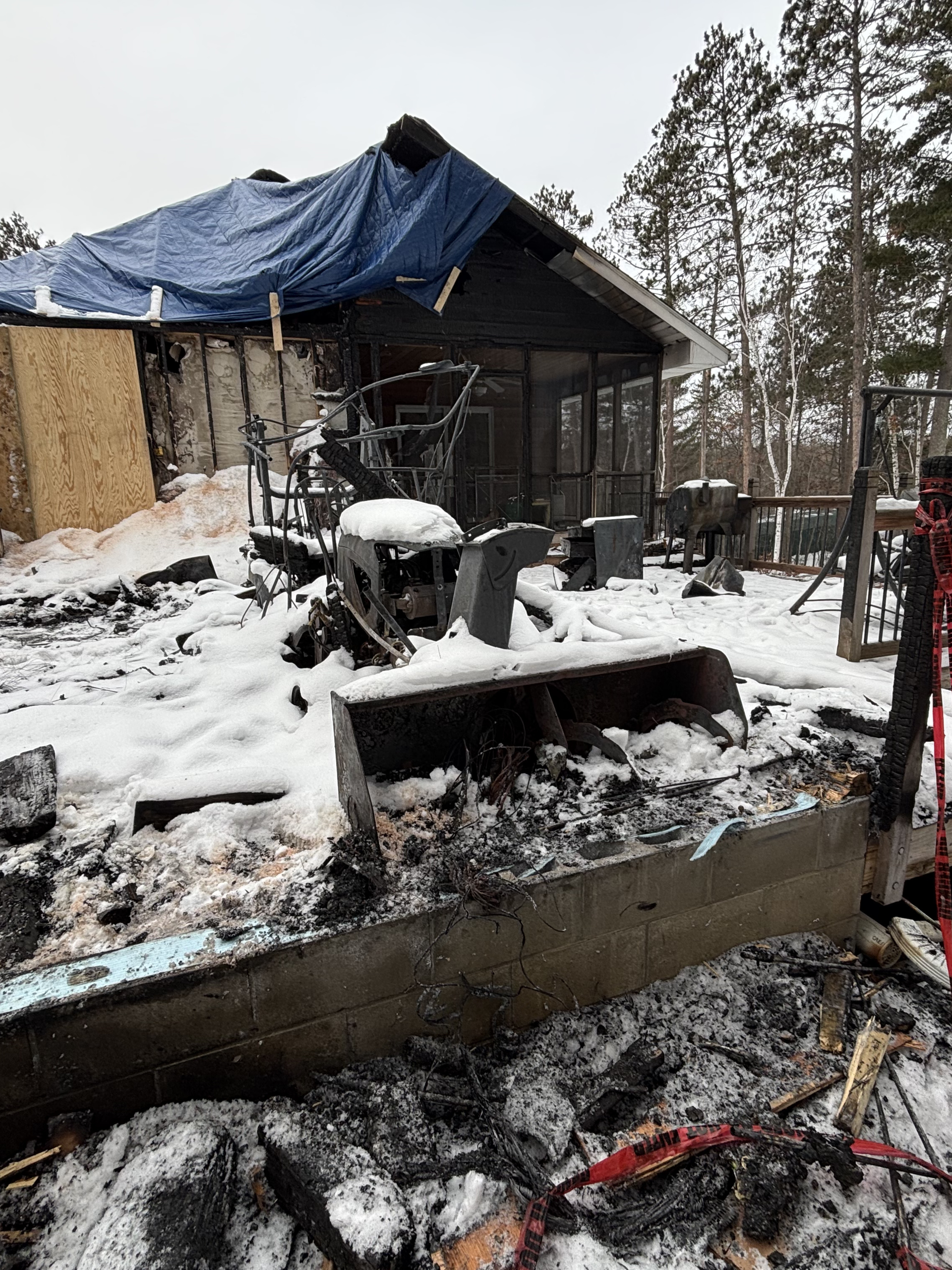 Snow-covered burned-out shed with collapsed roof and charred debris in a wooded yard