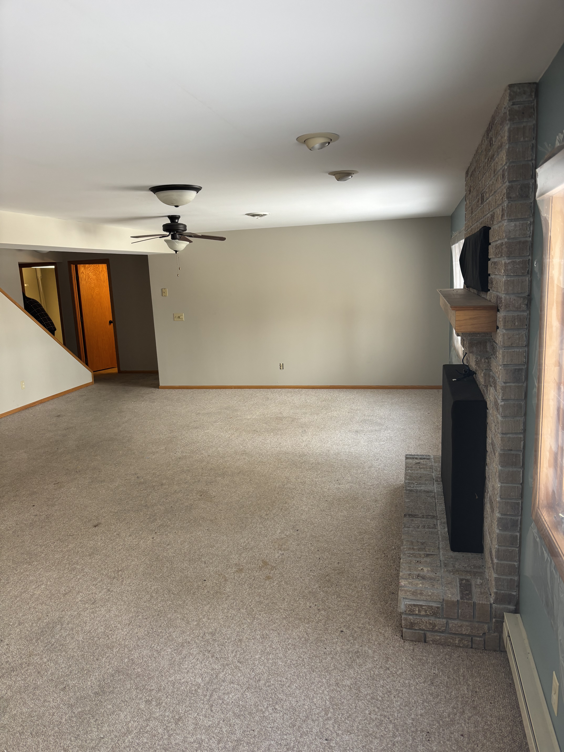 Empty carpeted living room with fireplace, ceiling fan, and hallway opening on the left.