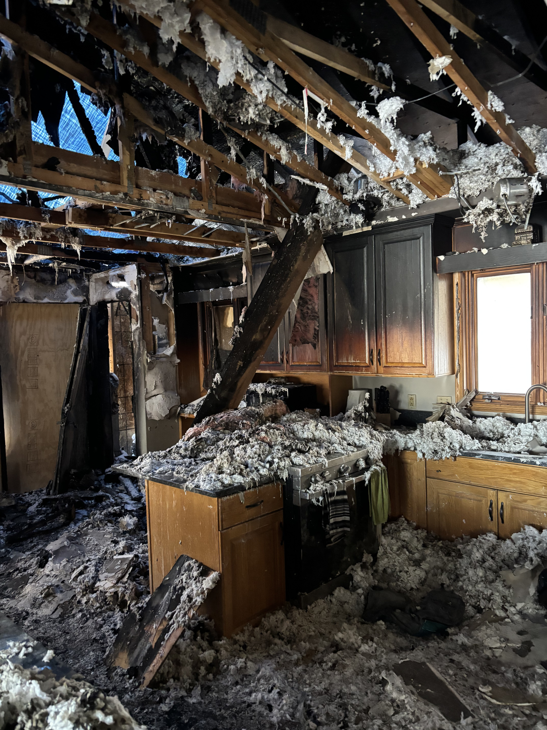 Burned kitchen interior with charred ceiling beams, collapsed debris, and blackened cabinets and walls