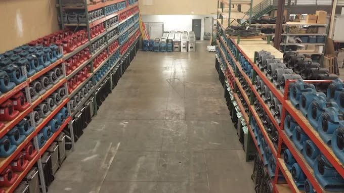 A warehouse aisle lined with tall industrial shelving stocked with numerous blue, red, and black air movers and fans.