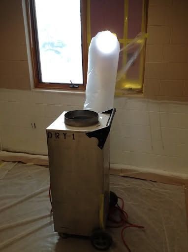 A stainless-steel industrial dehumidifier sits on a plastic-covered floor, venting air through a tube out of a window.