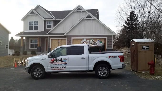 A white pickup truck with company branding parked in the driveway of a two-story suburban house next to a portable toilet.